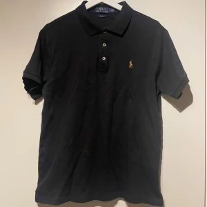Svart pikétröja från Ralph Lauren - Säljer en stilren svart pikétröja från Ralph Lauren i slim fit. Tröjan har korta ärmar och klassisk krage med knappar. Den ikoniska broderade loggan syns på bröstet. Perfekt för en avslappnad men ändå elegant look. Fint skick! Kolla gärna in min profil då jag rensar ut hemma och lägger ut mycket nu. Vid köp av flera artiklar kan rabatt ges!