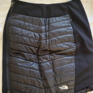 Svart kjol från The North Face - Snygg svart kjol från The North Face med quiltad design och dragkedja på sidan. Perfekt för kyligare dagar med sitt varma material. Stilren och funktionell för en aktiv livsstil.