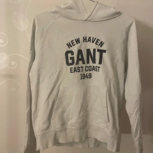 Vit hoodie från Gant - Säljer en vit hoodie från Gant med tryck 'New Haven East Coast 1949' i svart. Tröjan har en klassisk känguruficka och en bekväm huva. Perfekt för en avslappnad stil.