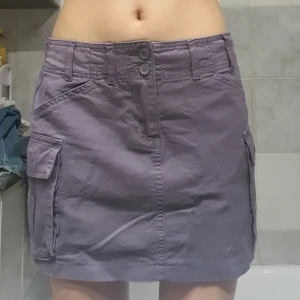 Lila kortkjol från Brandy Melville - ❗️Skriv innan köp❗️Lila kortkjol från Brandy Melville (Beverly Cargo Mini Skirt) med praktiska fickor på sidorna och dubbla knappar fram. Justerbara spännen baktill för bästa passform. Har använt ett fåtal ggr.