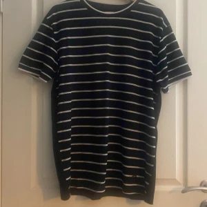 Svart och vit randig t-shirt från SAMS - Snygg svart t-shirt med vita ränder från SAMS. Den har en klassisk rund hals och korta ärmar. Perfekt för en avslappnad stil. Märkesdetalj på sidan ger en extra touch.