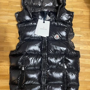 Svart väst från Moncler - Snygg svart dunväst från Moncler med avtagbar huva och dragkedja framtill. Västen har två praktiska fickor med dragkedja och Moncler-logga på bröstet. Perfekt för kyliga dagar. Storlek M men sitter som Xs-S