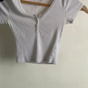 Vit ribbad topp från Gina Tricot - Säljer en vit ribbad topp från Gina Tricot med korta ärmar och knappar framtill. Perfekt för en stilren look. Toppen är croppad och har en rund halsringning.