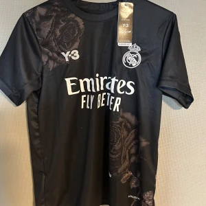 Real Madrid fotbolls tröja  - Snygg svart och ovanlig fotbollströja från Y3 med Real Madrid-logga och Emirates Fly Better-tryck. Tröjan har korta ärmar och ett subtilt blommönster. Perfekt för fotbollsfans som vill ha stil på planen. Funkar även utmärkt till vardags användning. Tröjan är även helt oanvänd och otroligt bra kvalitet.
