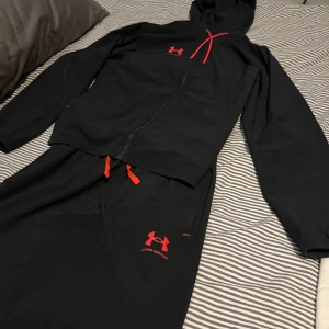 Svart träningsjacka från Under Armour - Säljer en svart träningsjacka från Under Armour med röd logga och detaljer. Litet hål längst ner på tröjan inget som syns