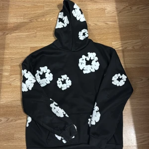 Svart hoodie med blommönster från Denim Tears - Säljer en äkta hoodie från Denim Tears med ett unikt vitt blommönster. Hoodien har en klassisk passform med långa ärmar och en bekväm huva. Perfekt för en stilren och avslappnad look. Den är knappt använd och är en bra skick