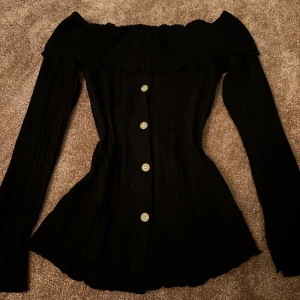 Svart offshoulder topp med knappar - Snygg svart offshoulder topp med långa ärmar och knappar framtill. Den har en ribbad struktur som ger en elegant look. Perfekt för en stilren outfit.