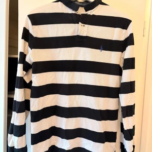 Randig pikétröja från Ralph Lauren - Säljer en klassisk randig pikétröja från Ralph Lauren i svart och vit. Tröjan har långa ärmar och en knappslå framtill. Perfekt för en stilren och avslappnad look.