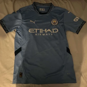 Manchester City home 24/25 fotbollströja  - Säljer en ljusblå fotbollströja från Puma med korta ärmar och svart text på kragen. Tröjan har ett tryck av ett klubbmärke och sponsorlogga på framsidan. Perfekt för fotbollsfans!