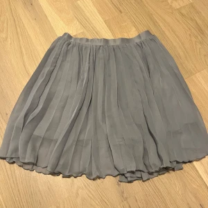 Grå kjol från H&M - Säljer en söt grå kjol från H&M. Kjolen har ett plisserat tyg som ger en fin volym och rörelse. Perfekt för både vardag och speciella tillfällen. Passar bra till en enkel topp eller en mer dressad blus.