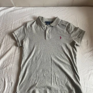 Grå pikétröja från Ralph Lauren - Den är L men är som M Säljer en stilren grå pikétröja från Ralph Lauren med en broderad rosa logotyp på bröstet. Tröjan är i slim fit och tillverkad av 100% bomull. Perfekt för en avslappnad men ändå snygg look.