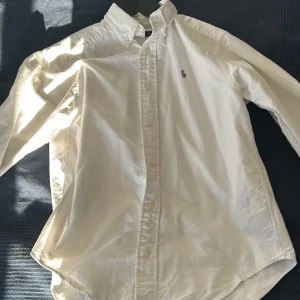 Vit skjorta från Ralph Lauren - Säljer en klassisk vit skjorta från Ralph Lauren med broderad logga på bröstet. Skjortan har långa ärmar och knappar framtill. Perfekt för en stilren look.