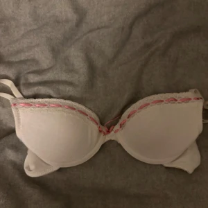 Vit bh med rosa detaljer - Säljer en söt vit bh med rosa spetsdetaljer och rosett mellan kuporna. Den har justerbara axelband för bästa passform. Perfekt för en bekväm och stilren look. I storlek 75 A-70 B