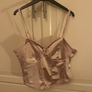Rosa satin korsett - Säljer en elegant rosa korsett i satin med justerbara axelband och en söt rosett framtill. Perfekt för en stilfull look.