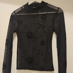 Svart mesh topp med sol- och månmönster - Säljer en svart mesh topp med ett coolt sol- och månmönster. Toppen har långa ärmar och en hög hals, perfekt för en unik stil. Passar bra till lager-på-lager outfits eller som statement piece. Strl S