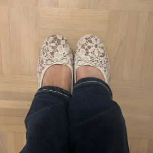 Vita spetsballerinaskor från H&M - Säljer ett par eleganta vita ballerinaskor från H&M med vacker spetsdesign och en söt rosett på ovansidan. Perfekta för en somrig look eller en speciell tillställning.