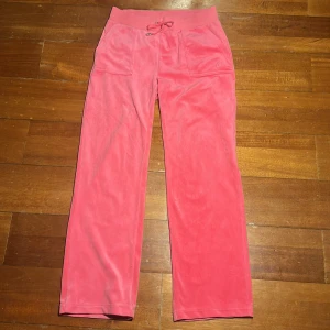 Rosa Juicy Couture mjukisbyxa - Snygg rosa JC mjukisbyxa med resår och snörning i midjan. Byxorna har två framfickor och två bakfickor med knappar. Perfekta för en avslappnad stil.