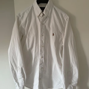 Vit skjorta från Ralph Lauren - Säljer en klassisk vit skjorta från Ralph Lauren i slim fit. Skjortan har långa ärmar och knappar framtill samt en broderad logotyp på bröstet. Perfekt för en stilren look.