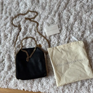 Svart axelväska från Stella McCartney - Snygg svart axelväska från Stella McCartney med guldfärgad kedja som axelrem och detaljer. Väskan har en stilren design och kommer med en dustbag. Perfekt för att ge en touch av lyx till din outfit.