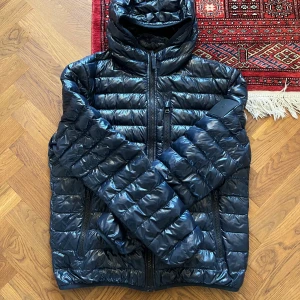 Blå dunjacka från Peak Performance - Säljer en snygg mörkblå dunjacka från Peak Performance. Jackan är glansig med  huva och dragkedja framtill. Perfekt för kalla vår och höstdagar och mildare vinterdagar. Använd i två vintrar, skick 8/10.  Passar M/L