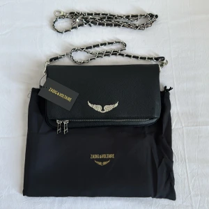 Svart axelväska från Zadig & Voltaire - Snygg svart axelväska från Zadig & Voltaire med silverdetaljer och kedjerem. Väskan har en elegant vingdetalj på framsidan och dragkedja för säker förvaring. Perfekt för att ge din outfit en stilfull touch.