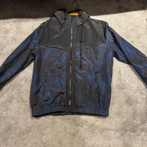 J Lindeberg vindjacka  - Riktig Snygg svart och blå vindjacka med ett coolt mönster riktigt fet i bilder när man använder blixt. Jackan har dragkedja framtill och justerbar huva. Perfekt för blåsiga dagar och ger en stilren look. 