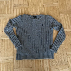 Grå stickad tröja från Ralph Lauren - Säljer en stilren grå stickad tröja från Ralph Lauren med kabelstickat mönster. Tröjan har långa ärmar och en diskret logga på bröstet. Perfekt för kyligare dagar.