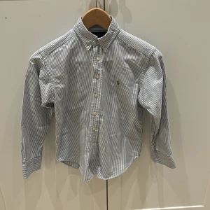 Randig skjorta från Ralph Lauren - Snygg randig skjorta från Ralph Lauren i blått och vitt. Skjortan har långa ärmar och knappar framtill. Perfekt för en stilren sommar look.