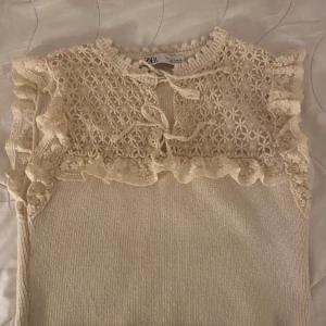 Vit spetsprydd topp från Zara - Säljer en söt vit topp från Zara med spetsdetaljer och volanger vid ärmarna. Toppen har en knytning framtill och är ärmlös, vilket gör den perfekt för varmare dagar. Den är tillverkad i ett mjukt material som känns skönt mot huden.
