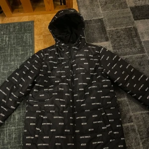 Svart pufferjacka från Vans - Säljer en svart pufferjacka från Vans med 'OFF THE WALL' tryck över hela jackan. Jackan har en dragkedja framtill och en huva för extra skydd mot väder. Perfekt för kyligare dagar. Den är aldrig använd. Varm vinterjacka