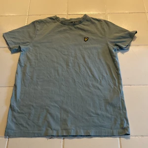Blå t-shirt från Lyle & Scott - Säljer en blå t-shirt från Lyle & Scott med ett broderat gult emblem på bröstet. T-shirten har en klassisk rund hals och korta ärmar. Perfekt för en avslappnad stil.