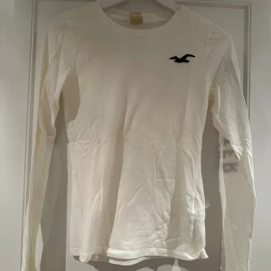 Vit långärmad tröja från Hollister - Säljer en vit långärmad tröja från Hollister med deras ikoniska logga på bröstet och texten 'Hollister' längs ärmen. Perfekt för en avslappnad stil. Tröjan är gjord i mjukt material och har en normal passform.