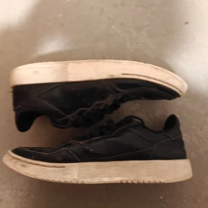Svarta sneakers - Säljer ett par svarta sneakers med snörning. Skorna har en vit sula och är tillverkade i syntetmaterial. Perfekta för vardagsbruk.