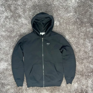 Cdg hoodie - Säljer en stilren svart hoodie från Comme des Garçons Play med dragkedja och det ikoniska hjärta-logotypen på bröstet. Perfekt för en casual look. M/L