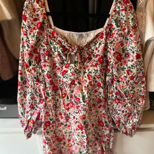 Blommig blus från NA-KD - Säljer en färgglad blommig blus från NA-KD med knytning framtill. Blusen har trekvartsärmar och en luftig passform, perfekt för vår och sommar.