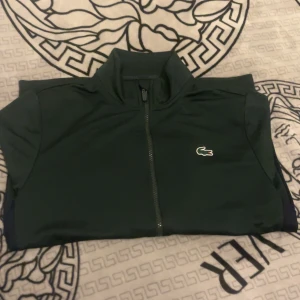 Grön jacka från Lacoste - Säljer en stilren grön dress från Lacoste med dragkedja och klassisk passform. Koftan har långa ärmar och det ikoniska krokodilmärket på bröstet. Perfekt för en sportig look.