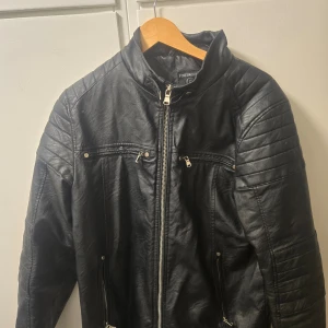 Svart läderjacka skinnimitation från Finesmekker mc biker med vaddering stl S/M - Snygg svart skinnjacka skinnimitation. från Finesmekker med dragkedja framtill och quiltade detaljer på axlarna. Jackan har flera praktiska fickor med dragkedjor och en stilren design som passar perfekt för en cool look samt vaddering på axlar och armbågar. Fint skick. Stl S/M.  Nypris 1999kr