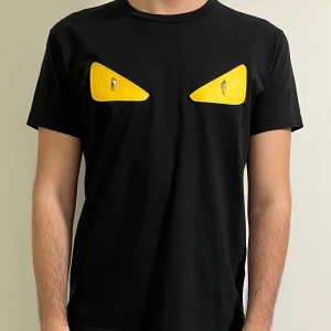 Svart t-shirt med gula detaljer från Fendi - Säljer en svart t-shirt från Fendi med coola gula ögon-detaljer på bröstet. T-shirten är använd max 5 gånger och köpte den i Milano för något år sedan och säljer jag den då den inte används. T-shirten är kortärmad och gjord i mjukt material för en bekväm passform, storleken är XL men funkar som L. 