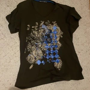 Svart t-shirt med emo mönster - Snygg svart baggy t-shirt med ett unikt blått och beige mönster framtill. T-shirten har en v-ringad hals och korta ärmar, perfekt för en y2k stil. Passar bra till jeans eller shorts för en cool look.