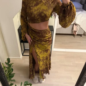 Gul och brun långklänning med slits - Trendig brun, guldig långklänning med ett unikt mönster. Klänningen har långa ärmar och en snygg slits framtill. Perfekt för att sticka ut. Aldrig använd och prislapp är kvar! 