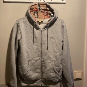 Grå hoodie från Burberry - Säljer en stilren grå hoodie från Burberry med dragkedja och broderad logga på bröstet. Den har en klassisk huva med snörning och en mjuk insida. Perfekt för en avslappnad look.