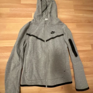 Grå hoodie från Nike - Säljer en grå hoodie från Nike med dragkedja och svart detaljering. Den har en huva och långa ärmar med en svart rand på ärmen. Perfekt för en sportig look eller avslappnad stil.