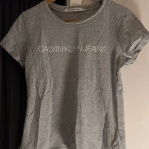 Grå t-shirt från Calvin Klein Jeans - Säljer en stilren grå t-shirt från Calvin Klein Jeans med logotypen tryckt på bröstet. T-shirten har en rund halsringning och korta ärmar. Perfekt för en avslappnad look att bara gosa i hemma eller klä upp med accessoarer💕