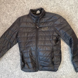 Svart pufferjacka från Emporio Armani - Säljer en svart pufferjacka från Emporio Armani EA7. Jackan har en quiltad design med dragkedja framtill och två sidofickor. Perfekt för kyligare dagar med sin stilrena look och bekväma passform.