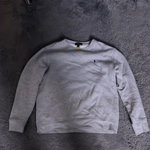 Grå tröja från Polo Ralph Lauren - Säljer en stilren grå tröja från Polo Ralph Lauren med deras ikoniska logga broderad på bröstet. Tröjan har långa ärmar och en klassisk rund halsringning. Perfekt för en avslappnad look. Xl i barn storlek