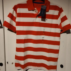 Röd och vit randig pikétröja från Tommy Hilfiger - Säljer en snygg röd och vit randig pikétröja från Tommy Hilfiger. Tröjan har en klassisk krage och knappar framtill. Perfekt för en avslappnad stil med en touch av elegans. Passar både till jeans och shorts! Barnstorlek 12-14 år typ 158-164