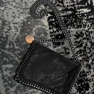 Svart axelväska med kedjedetaljer - Snygg svart axelväska med glittrig yta och kedjedetaljer längs kanterna. Väskan har en praktisk dragkedja på baksidan och en kedja som axelrem. Perfekt för att ge din outfit en edgy touch.