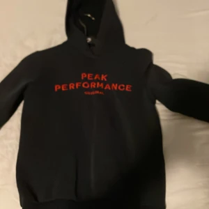 Svart hoodie från Peak Performance - Säljer en svart hoodie från Peak Performance med röd text på bröstet. Tröjan har en klassisk design med huva och långa ärmar. Perfekt för en avslappnad stil. OBS! Trasig vid handledens slut på tröjan