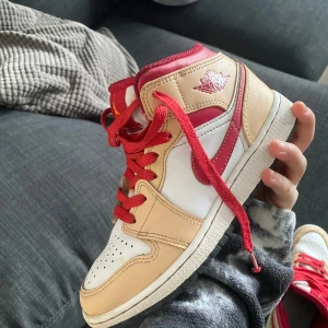 Nike Air Jordans i beige och rött - Snygga Nike Air Jordans i beige och rött med vita detaljer. Skorna har röd snörning och en klassisk design med perforerad tå. Perfekta för dig som vill ha en stilren och sportig look.