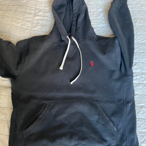 Svart hoodie från Ralph Lauren - Säljer en stilren svart hoodie från Ralph Lauren med en liten röd logga på bröstet. Tröjan har en klassisk känguruficka och justerbar huva med vita snören. Nypris 2295